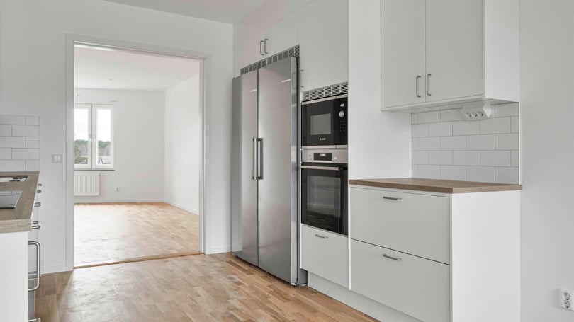 Modern kök med vita skåpluckor, rostfria vitvaror, mikrovågsugn, ugn och kylskåp. Öppet utrymme med trägolv leder till ett ljusfyllt rum i bakgrunden