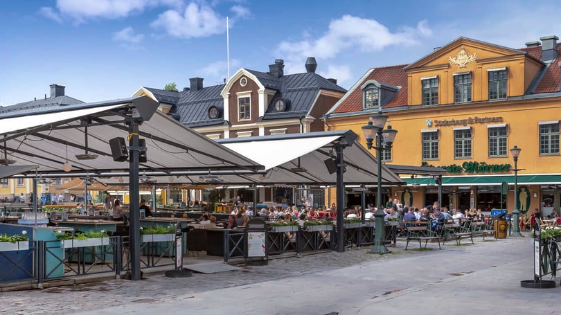 Uteservering på ett torg med människor och färgglada byggnader i bakgrunden