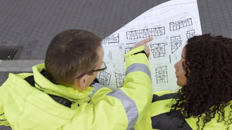 Två personer i gula reflexjackor studerar en ritning med bostadslayouter