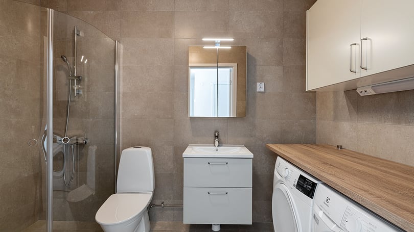 Modernt tvättutrymme med dusch, toalett, handfat och tvättmaskin. Beige kakel och förvaringsskåp