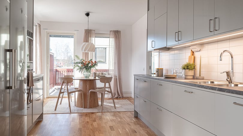Ljust kök med matplats, grå köksluckor, träbord och stora fönster med utgång till balkong