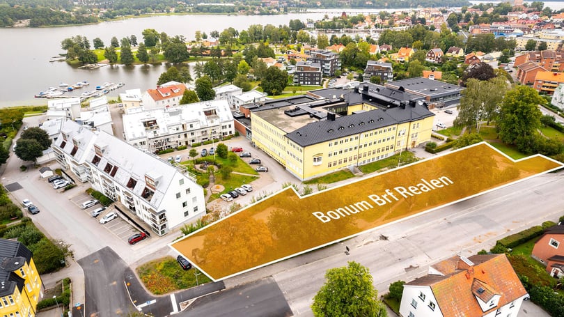Flygfoto över bostadskvarter vid vatten med flerbostadshus, vägar och båtbrygga. Markering med texten Bonum Brf Rejlen syns på en tom yta