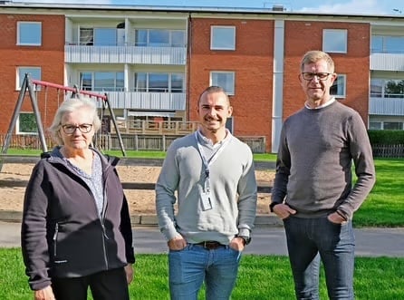 Tre personer står framför en lekplats med gungor, gräs och ett flerfamiljshus i bakgrunden