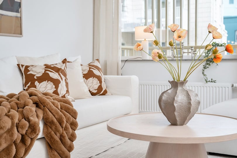 Ljus vardagsrum med vit soffa, bruna mönstrade kuddar, beige filt och blomvas med orange och vita blommor på ett runt träbord