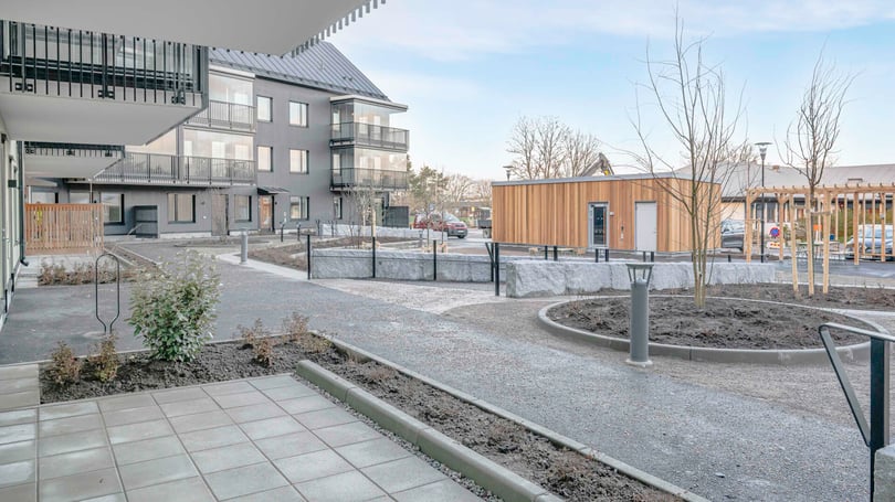 Gårdsplan med moderna flerfamiljshus, nyplanterade träd, och en parkeringsplats i bakgrunden under en klar himmel