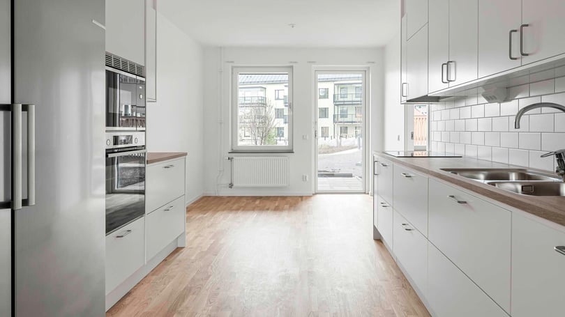 Modernt kök med vita skåp, inbyggd ugn och mikrovågsugn, träparkettgolv samt utsikt mot en innergård genom ett stort fönster och altandörr