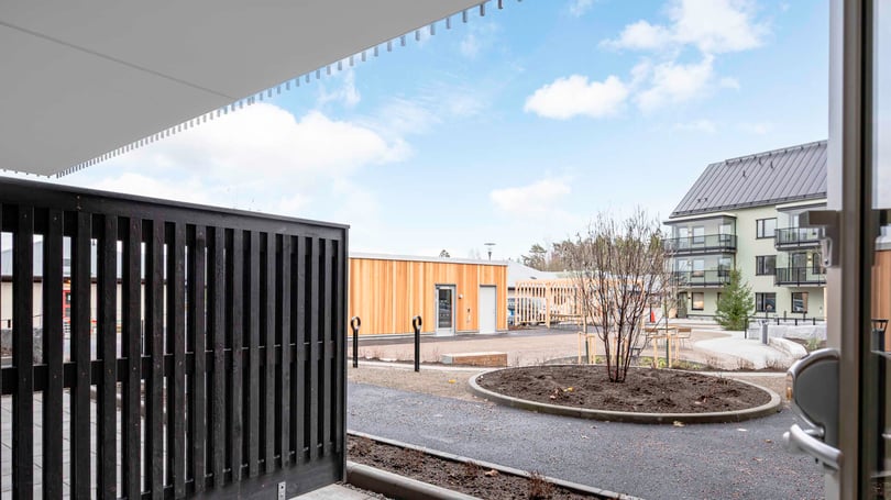 Utsikt från en altan mot en innergård med moderna byggnader, asfaltvägar och planterade buskar under en blå himmel med moln
