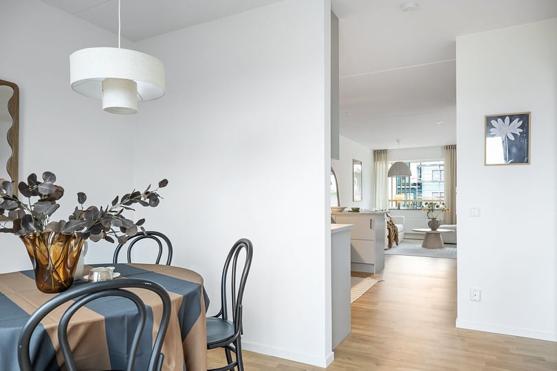 Interiör av ett modernt hem med matplats, inkluderande ett runt bord med bordduk, svarta stolar och en vas med dekorativa växter. Bakgrunden visar ett vardagsrum med ljusa möbler och stora fönster
