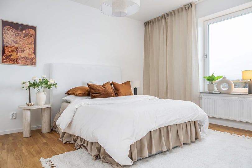 Sovrum med en stor säng klädd i vitt och beige sängkläder, vita väggar, ett fönster med beige gardin, och en träpanel på golvet. En nattduksbord med blommor och en tavla på väggen