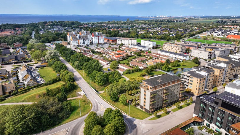Flygfoto över ett bostadsområde med byggnader, grönområden och vägar, samt hav och horisont i bakgrunden