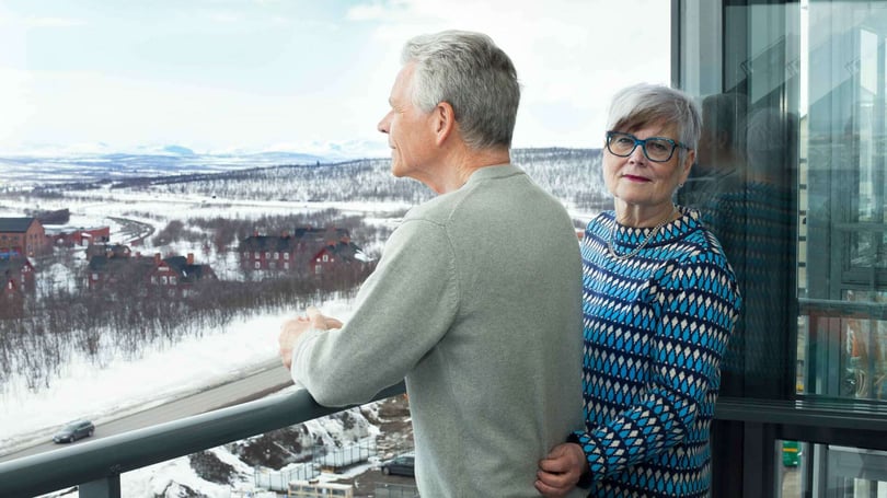Två äldre personer står på en balkong och blickar ut över ett snötäckt landskap