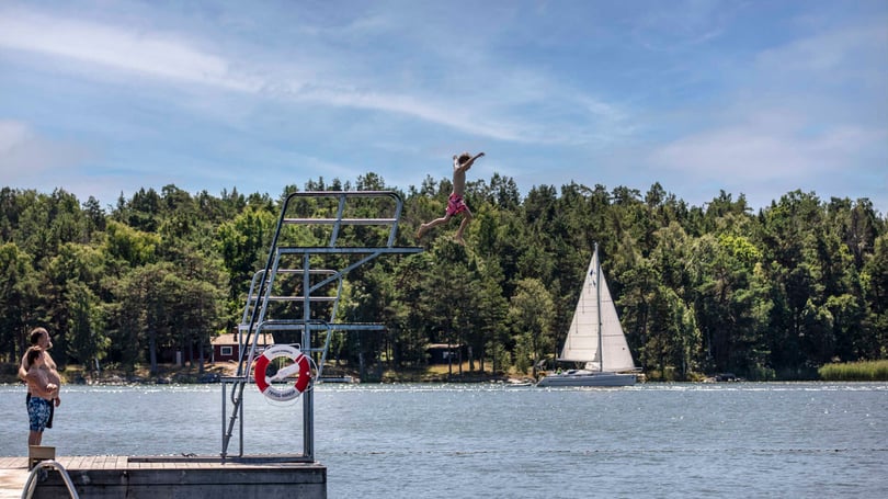 Person som hoppar från ett hopptorn vid en sjö, med segelbåt och skog i bakgrunden