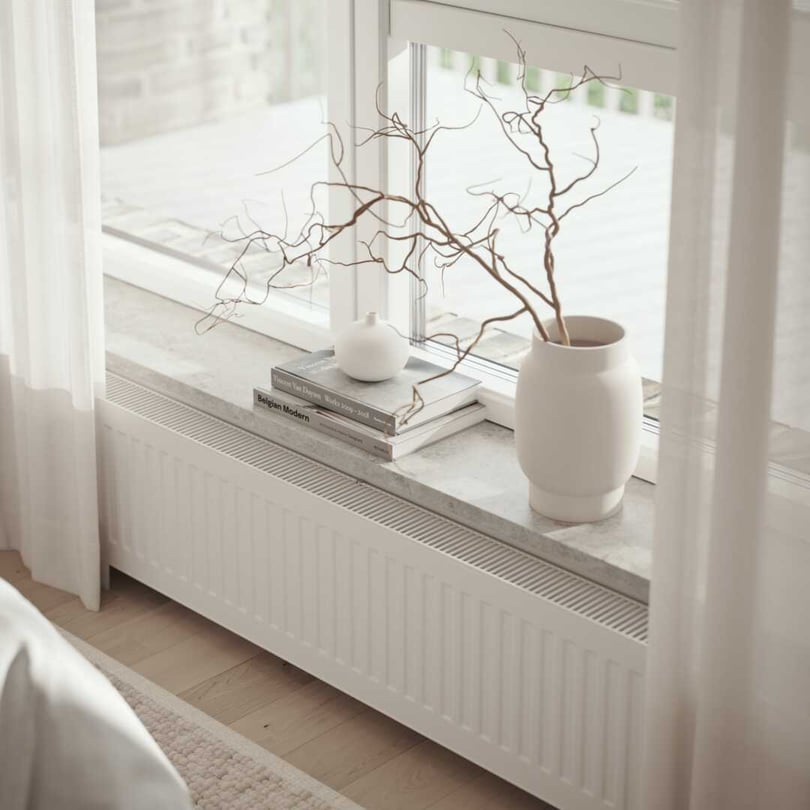 Ett fönster med vita gardiner, en marmorfönsterbräda dekorerad med en vit vas med torra grenar, en vit kruka och böcker