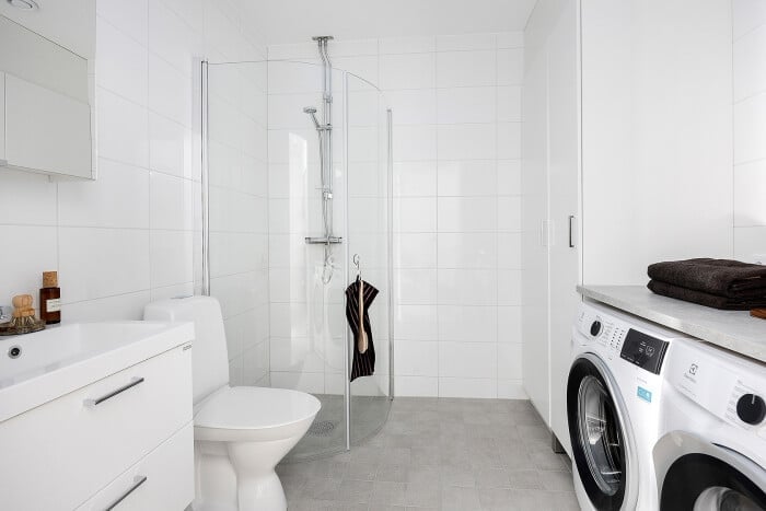 Modernt badrum med vit kakelvägg, dusch med glasvägg, tvättmaskin, toalett och handfat med spegelskåp