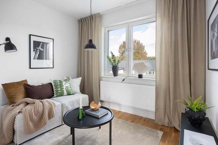 Vardagsrum med ljusa väggar, en vit soffa med kuddar och filt, ett runt soffbord, gröna växter och stora fönster med beige gardiner