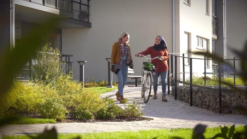 Två kvinnor går utanför ett bostadshus, en leder en cykel