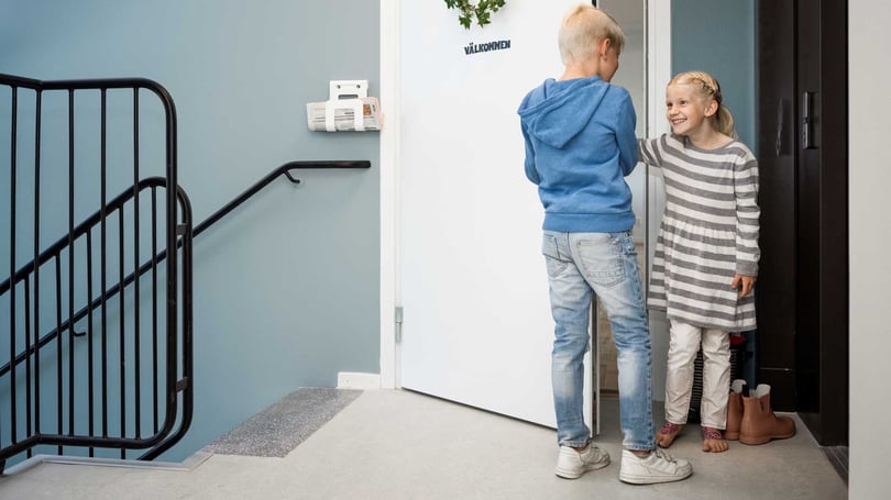 Två barn står vid en vit dörr i en trappuppgång och tittar in i en lägenhet