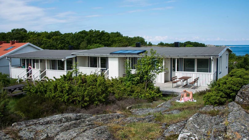 Fritidshus vid havet med altan och grönska, samt en person som ligger på en klippa i förgrunden