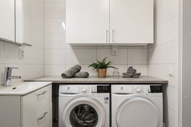 Tvättstuga med tvättmaskin och torktumlare under en bänkskiva, vitt kaklade väggar och handfat