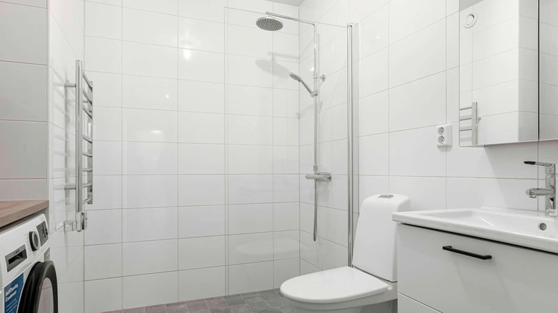 Badrum med vitkaklade väggar, dusch med glasvägg, toalett, tvättmaskin och vit handfat med spegelskåp