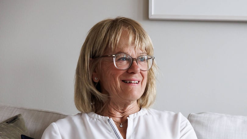 Äldre kvinna med glasögon som sitter i en soffa och ler, med ljus inomhusmiljö i bakgrunden