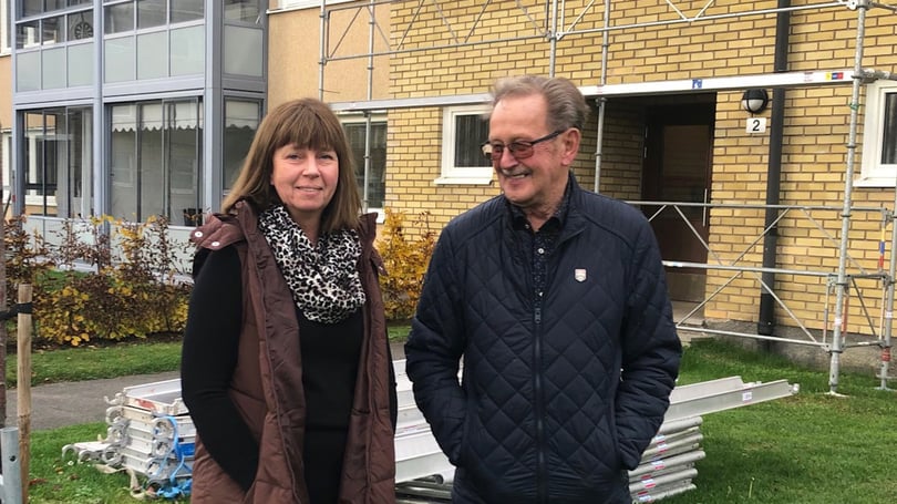 Två personer står utomhus framför en gul tegelbyggnad med byggnadsställning
