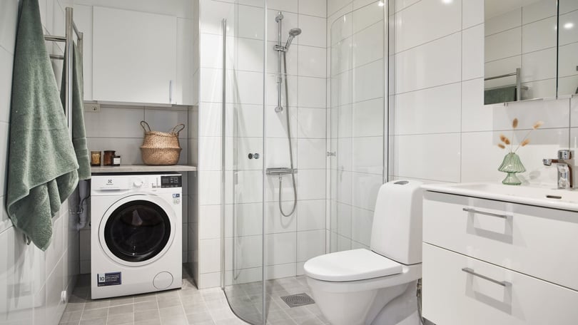 Modernt badrum med vit kakelvägg, glasdusch, tvättmaskin, vitt skåp och toalett