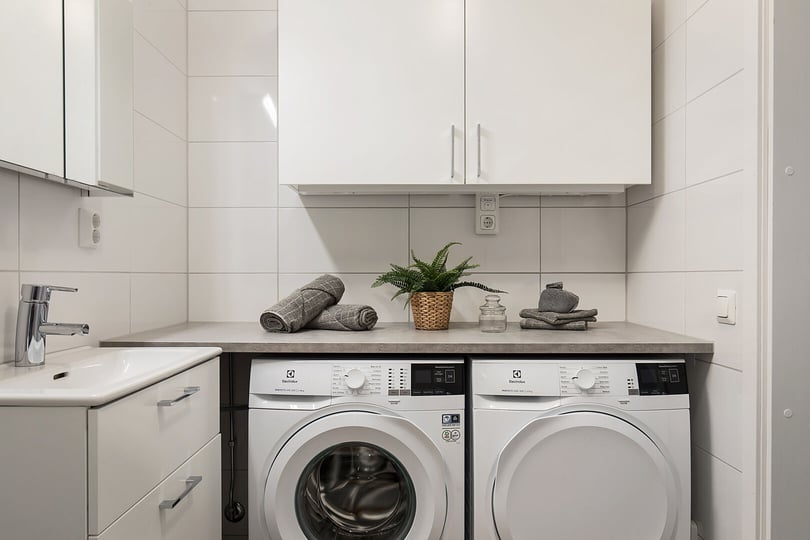 Tvättstuga med tvättmaskin och torktumlare under en bänkskiva, vitt kaklade väggar och handfat