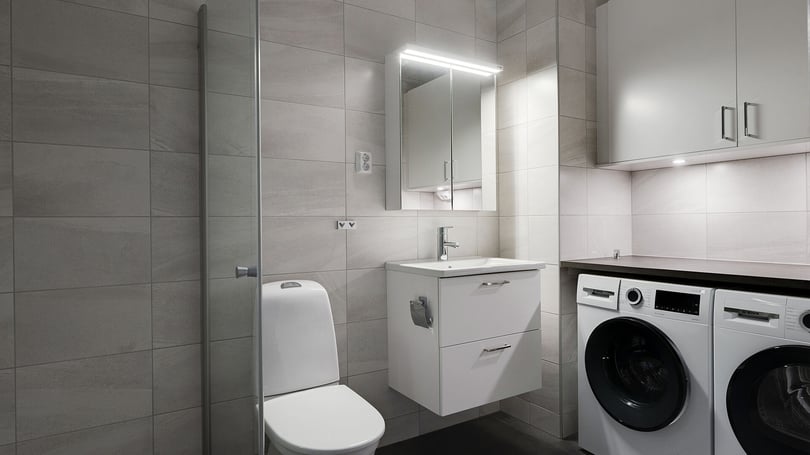 Badrum med grå kakelväggar, kommod med handfat, spegelskåp, duschvägg och tvättmaskin samt torktumlare