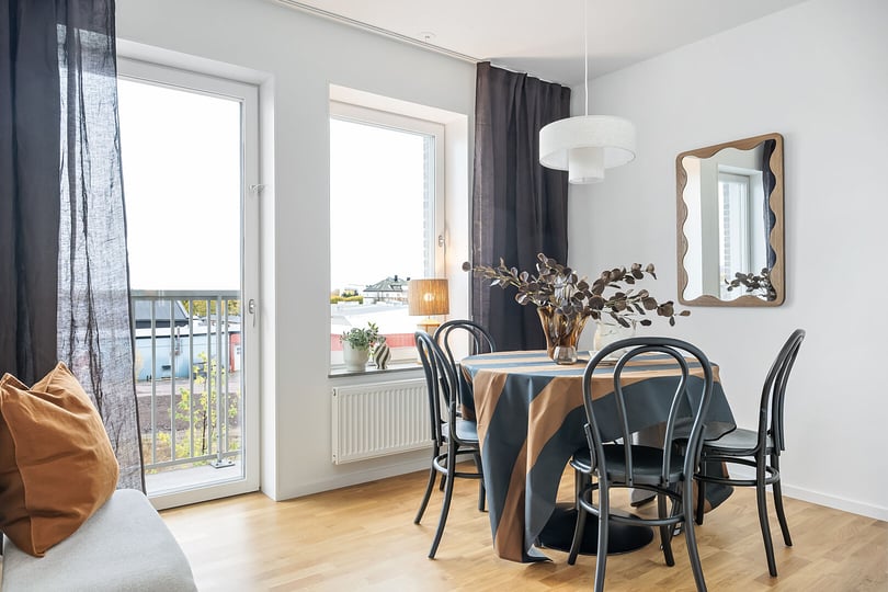 Ljust matrum med stora fönster, svart matbord med svarta stolar, randig bordsduk och dekorativ spegel på väggen
