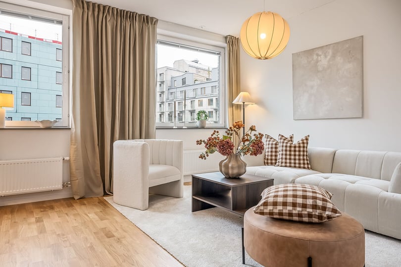 Modernt vardagsrum med ljus inredning, beige soffa, schackrutiga kuddar och ett bord dekorerat med blommor. Stora fönster med utsikt över stadens byggnader och en rund pendellampa i taket