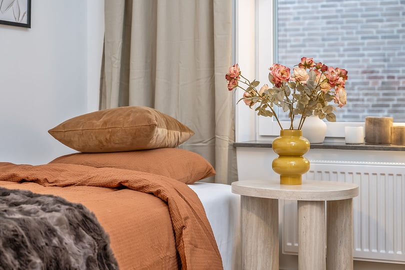 Sovrum med en orange sängklädsel, en beige kudde och ett sidobord med en gul vas fylld med rosor. Fönster med ljusinsläpp och en synlig tegelvägg utomhus