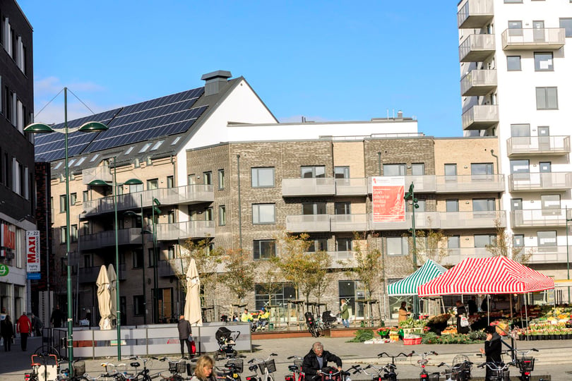 Bostadshus med balkonger och solpaneler vid ett torg med marknadsstånd