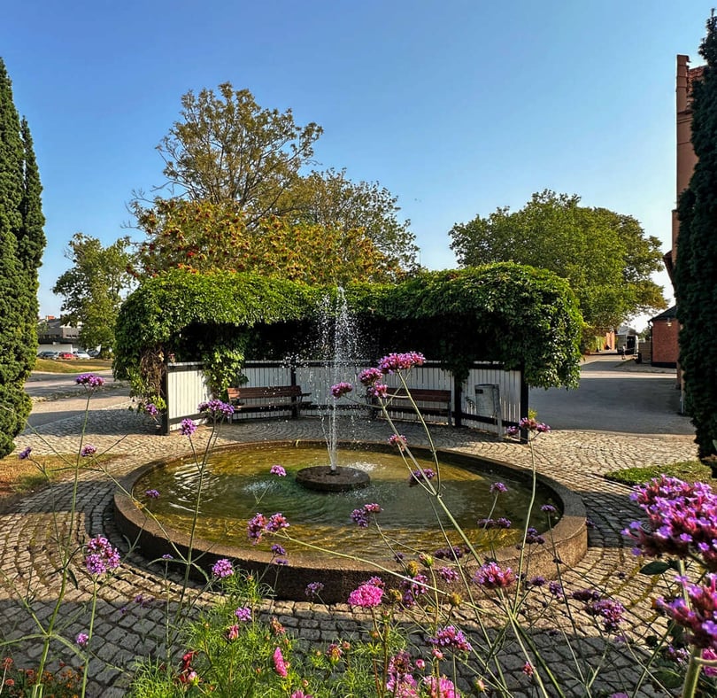 Brunn med fontän i en grönskande innergård omgiven av blommor och kullersten