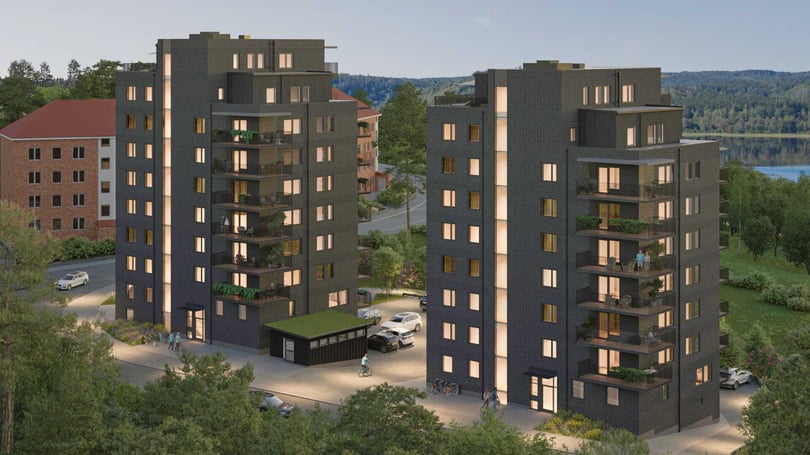 Två moderna lägenhetsbyggnader med mörk fasad och flera våningar, omgivna av grönska och parkerade bilar, med en sjö och skog i bakgrunden