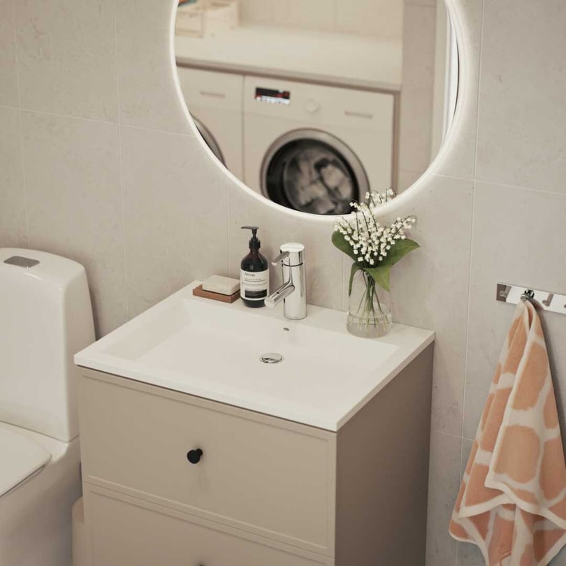 Modernt badrum med vit toalett, beige handfatsskåp, rund spegel med belysning, blomvas och tvättområde synligt i bakgrunden