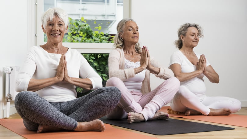 Tre personer som sitter på yogamattor i en meditationsställning inomhus, med ett fönster och grönska i bakgrunden