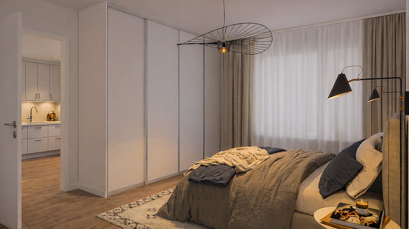 Sovrum med en dubbelsäng, beige gardiner, taklampa och golvlampa. En dörr leder till ett kök med ljusa skåp och träfärgat golv