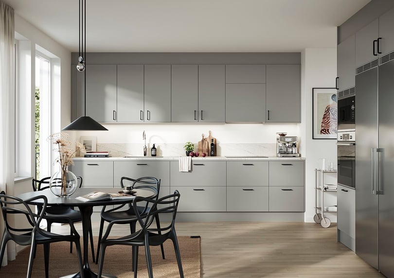 Modernt kök med gråa skåpluckor, vit bänkskiva och rostfria vitvaror. Matbord med svarta stolar står på en beige matta nära stora fönster