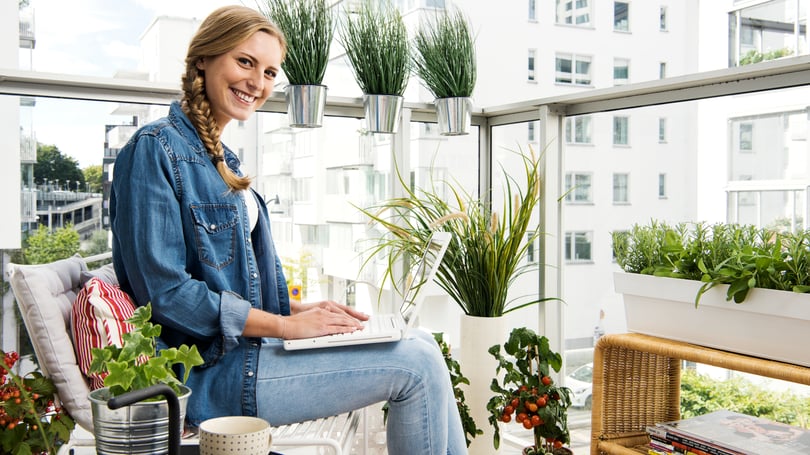 Kvinna som sitter på en inglasad balkong med laptop, omgiven av gröna växter