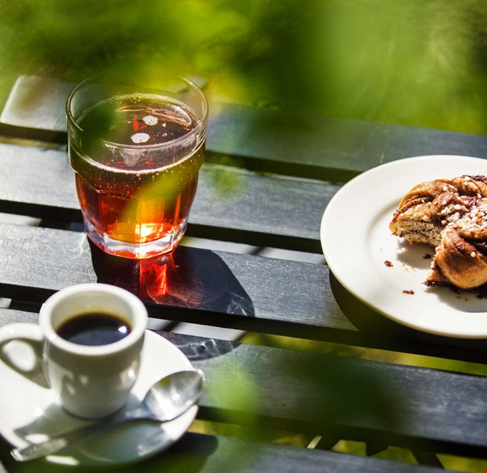 Ett glas saft, en kopp kaffe och en bulle på ett bord i solljuset
