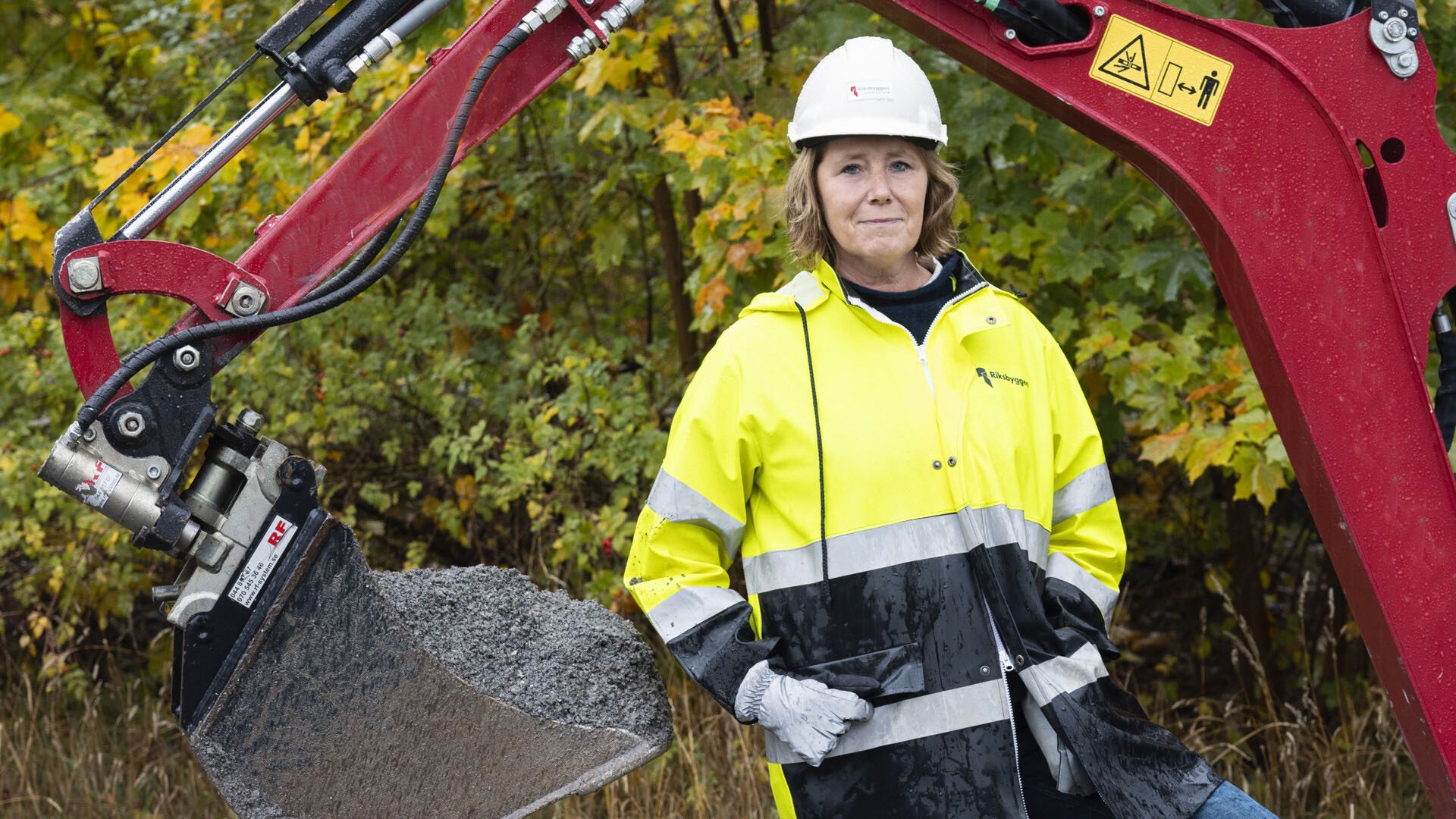 Riksbyggens vd Johanna Frelin iklädd en gul arbetsjacka stående framför en röd grävskopa på en byggplats